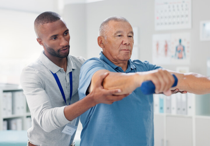 A physical therapist in Apopka, Florida assists an older man in a light blue shirt with arm exercises using small blue dumbbells in a clinical setting.