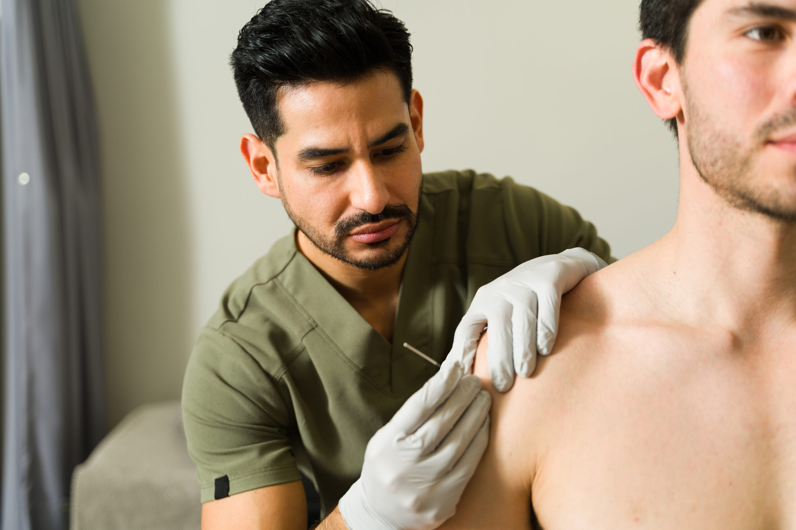 A healthcare professional in scrubs and gloves inserts an acupuncture needle into a patients bare shoulder.