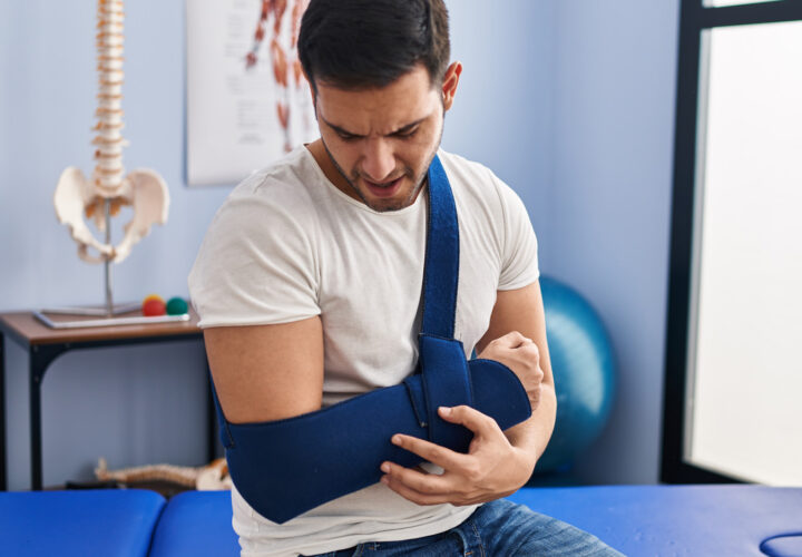 A man sits on a medical bench, his left arm in a blue sling, appearing to be in pain—an example of Post-Surgical Rehabilitation with an anatomy poster on the wall behind him.