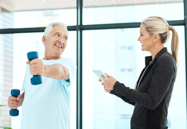 An older man lifts dumbbells while a woman in athletic wear, specializing in Post-Surgical Rehabilitation, holds a tablet and smiles at him in a PT clinic in Apopka, FL.