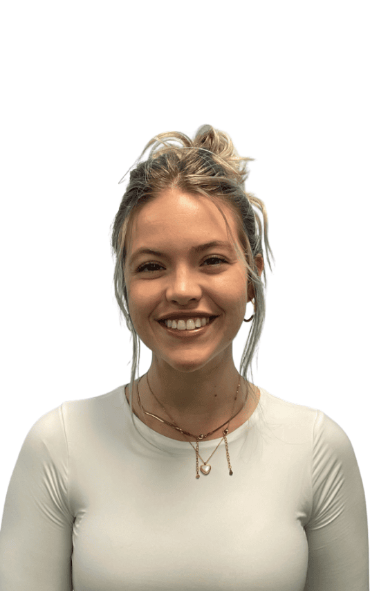 A young woman with blonde hair in a messy bun, wearing a white long-sleeve shirt and layered gold necklaces, smiles at the camera against a plain white background.