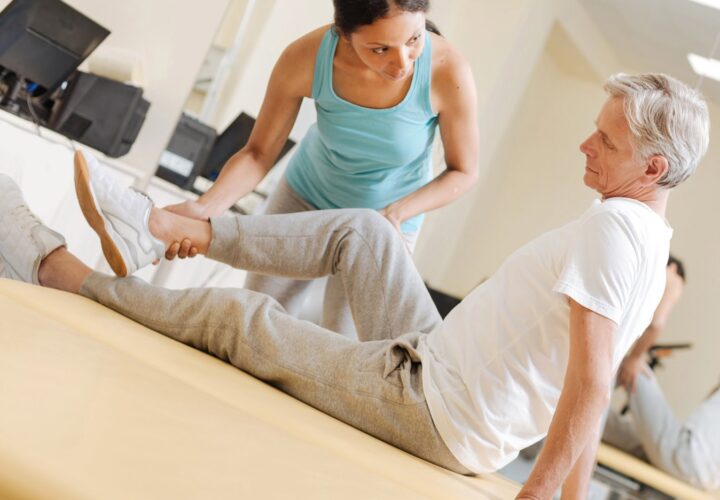 A woman assists an older man with a leg stretch on a treatment table during Post-Surgical Rehabilitation in an Ocoee physical therapy clinic.