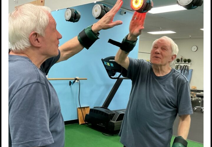 An older man wearing wrist weights touches a glowing light on a mirrored wall in a gym or physical therapy setting.