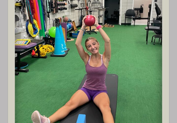 Woman in athletic wear sits on a mat in a gym, holding a red ball overhead with both hands, legs extended, exercise props visible around her.