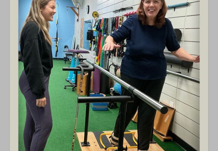 Two women are in a physical therapy room; one is smiling while balancing on a platform with support bars, and the other stands nearby observing.