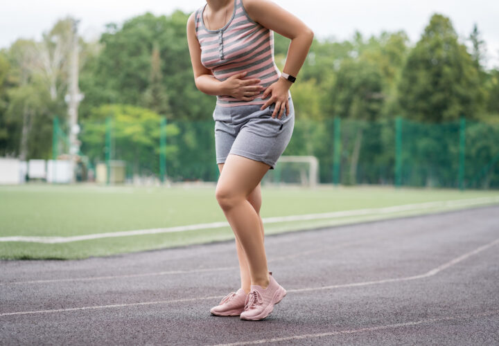 A person standing on a running track, holding their stomach and side, appearing to be in discomfort or pain. Trees and a soccer field are visible in the background.