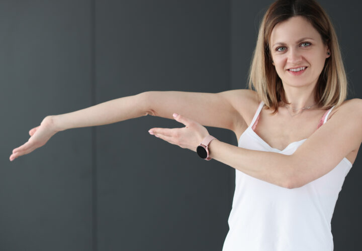 Woman in a white tank top stretches her right arm across her chest, smiling, against a plain dark background.